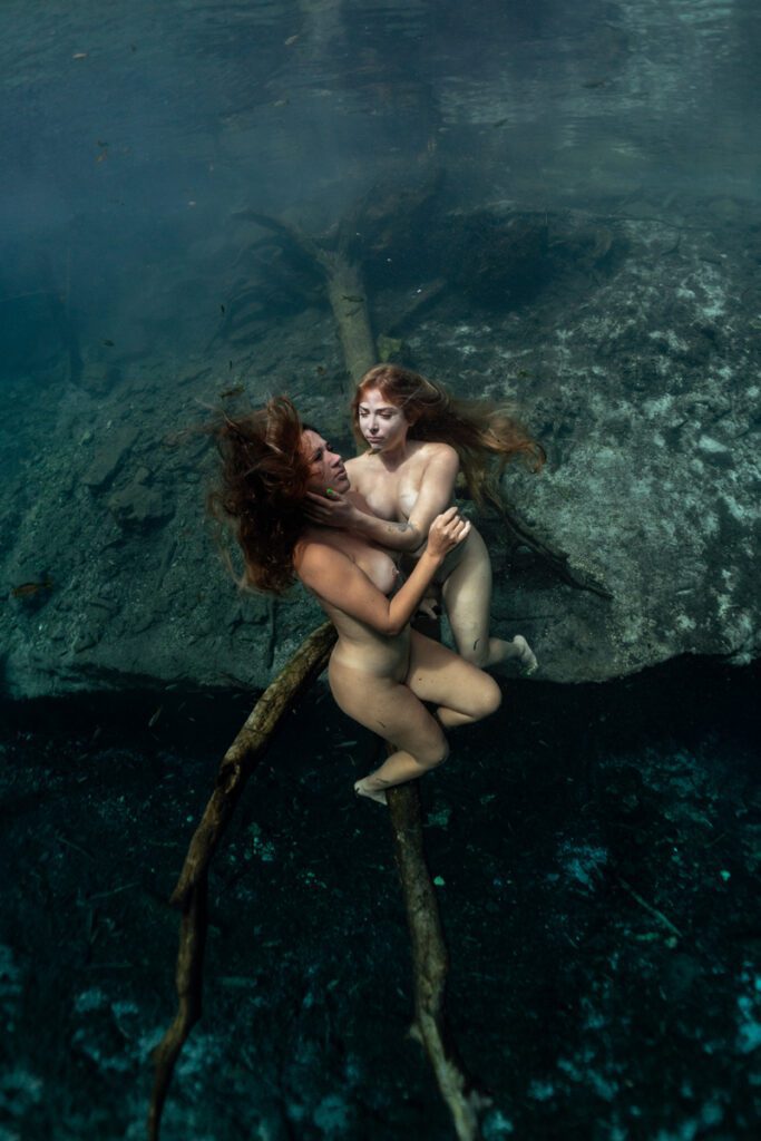 two women underwater in a cenote sitting on the branch of a tree. One touches the other's face.