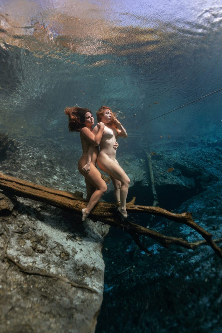 two beautiful girls naked and posing standing and hugging on the branch of a tree, posing underwater in cenote.