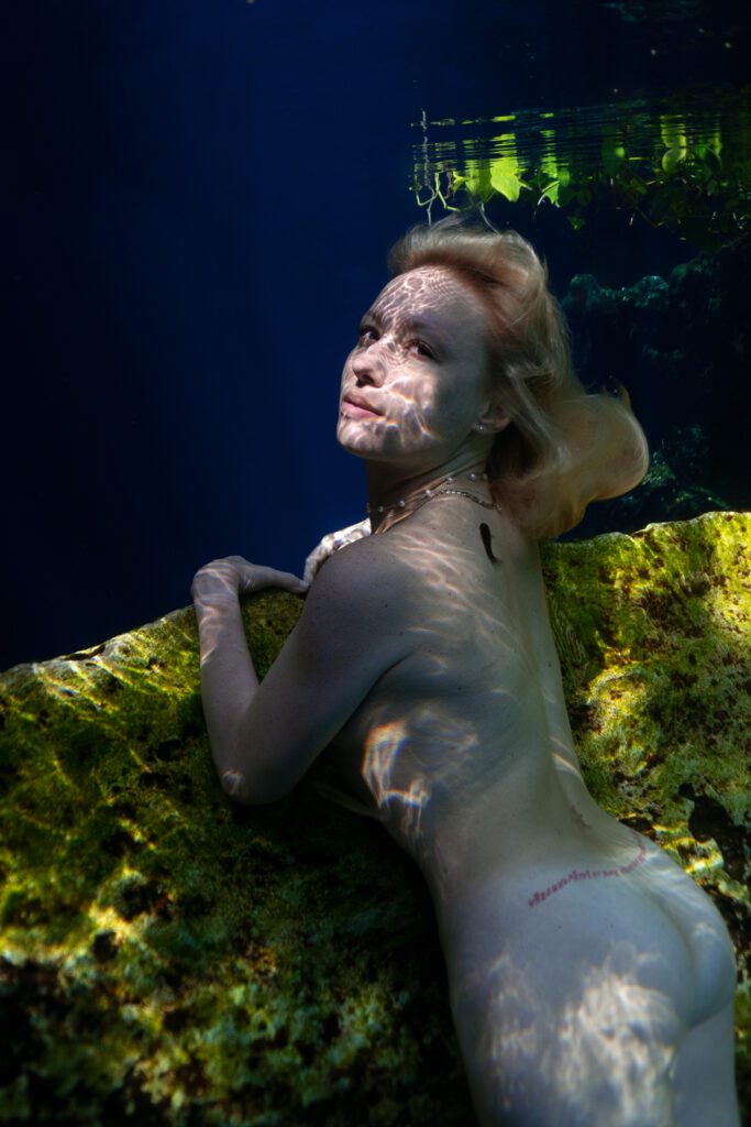 Medium shot of a blonde woman from behind turning around and looking at the camera naked underwater in a cenote in Mexico.
