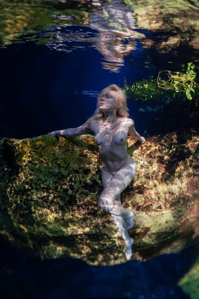 A blonde woman poses naked on a yellow rock underwater in a cenote. She is facing the front with her two arms resting on the rock.