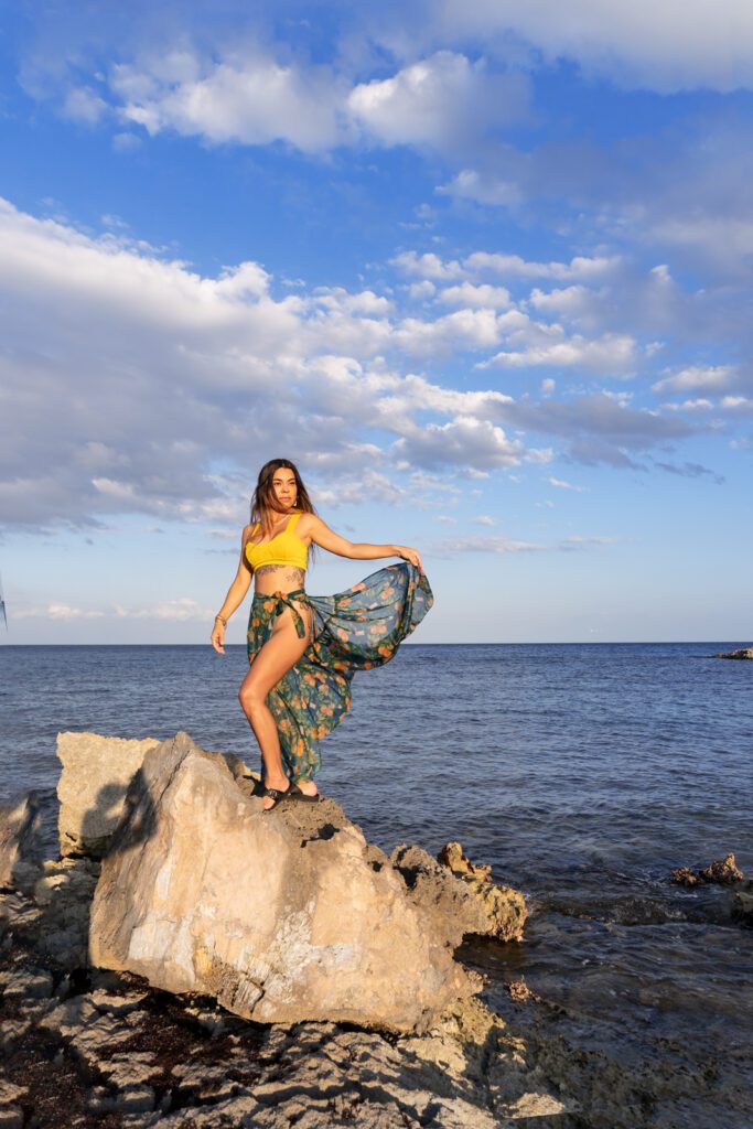 Scenic view of an individual standing on a rock by the sea, wearing a long skirt. The photo captures the full body, and the background showcases the sky and the vast expanse of the sea, creating a picturesque coastal scene.