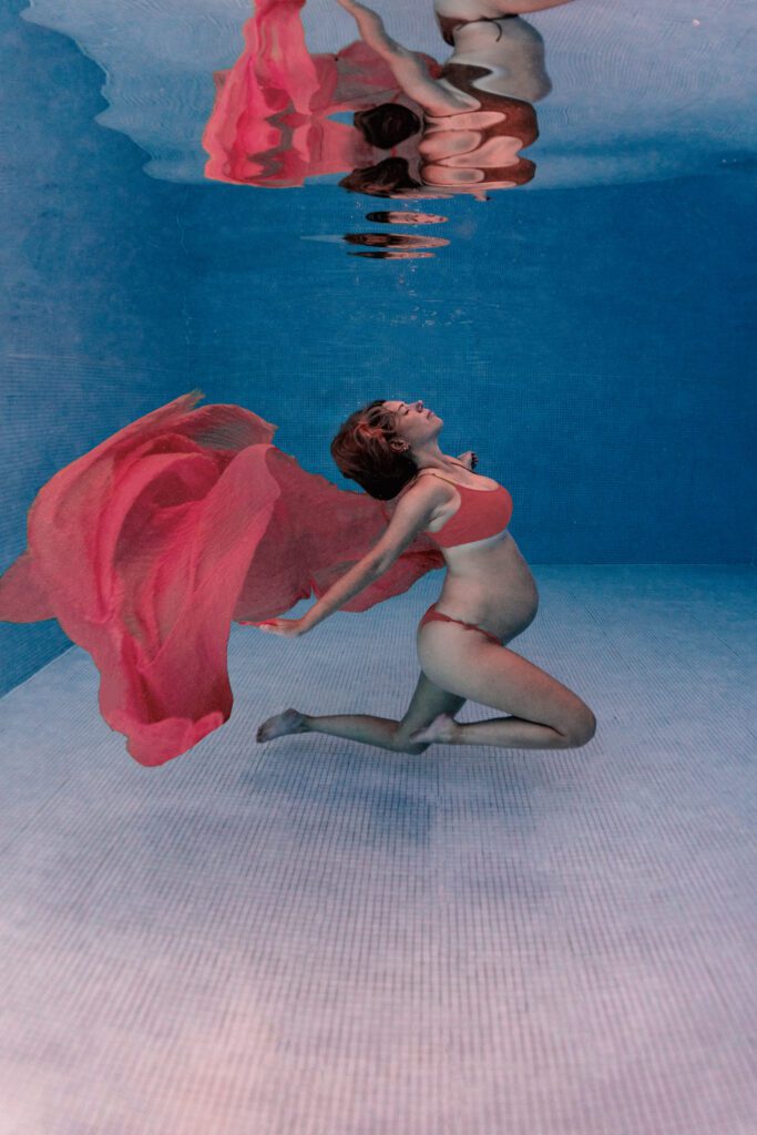 a pregnant girl posing underwater with a bikini and a pink cloth that is moving behind her.