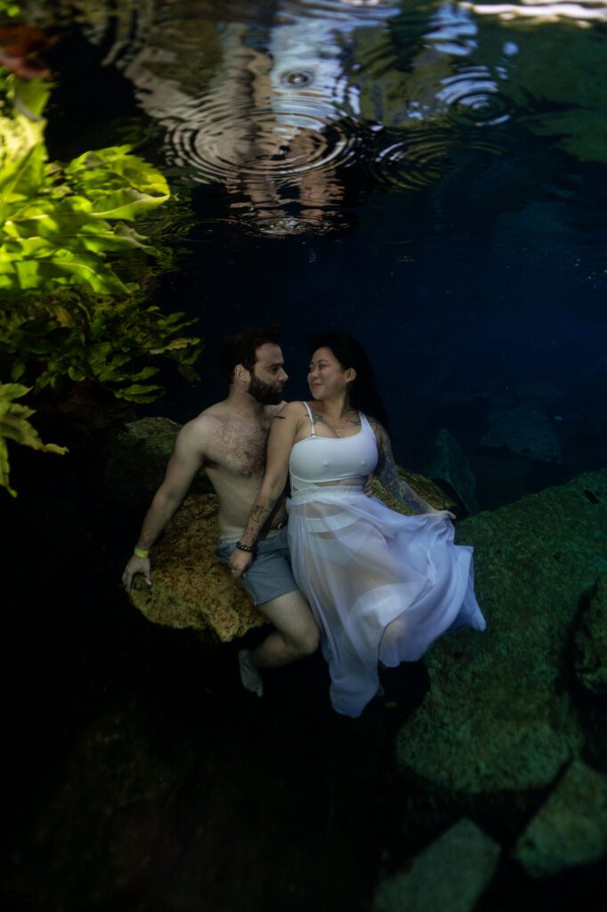 a couple in love is sitting on a rock underwater looking at each other, there are green underwater plants around them