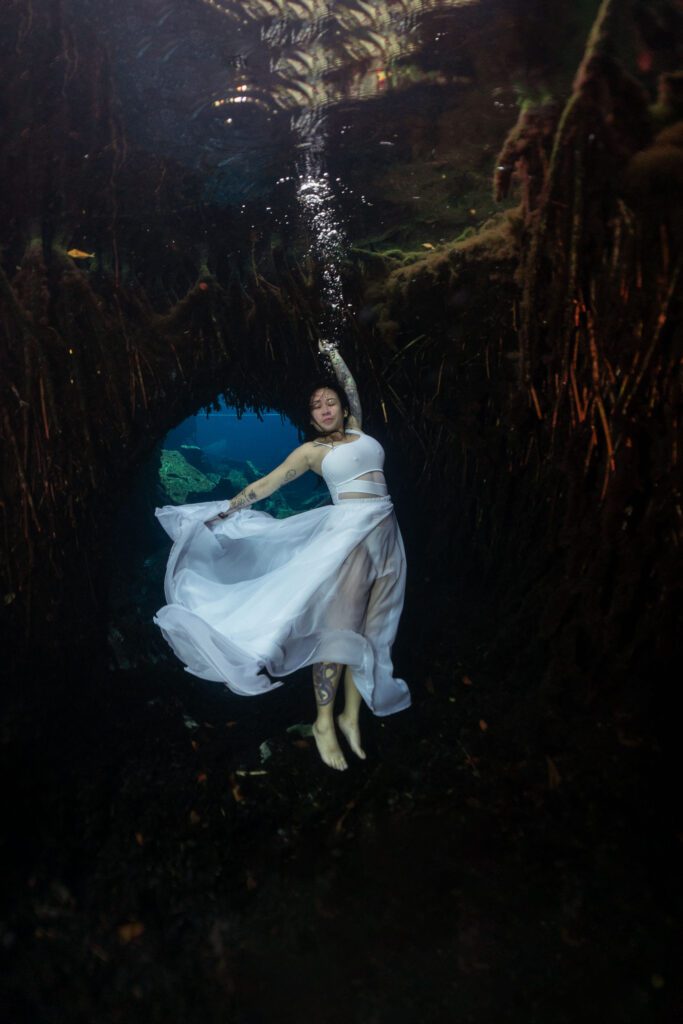 An Asian girl poses underwater with a white dress, in the background you can see an underwater cave inside a cenote.