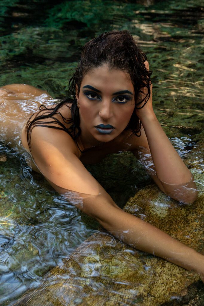 A beautiful girl lying naked in the water in a cenote, face down with her hand on her head, looks at the camera.