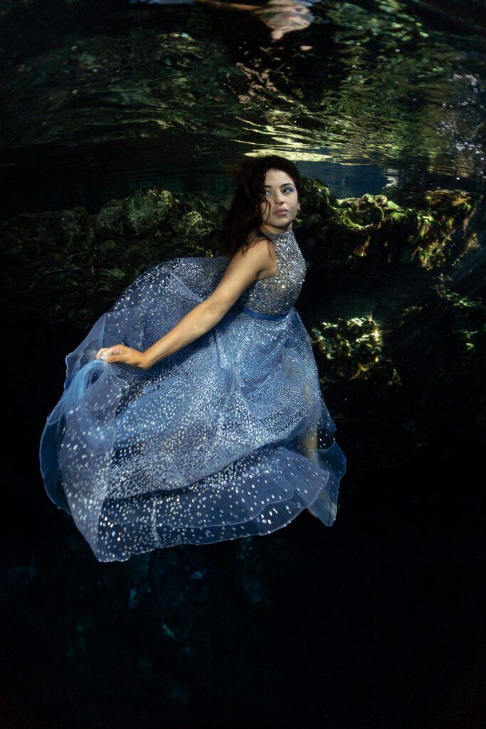 a beautiful woman poses underwater in a cenote with a light blue and bright dress