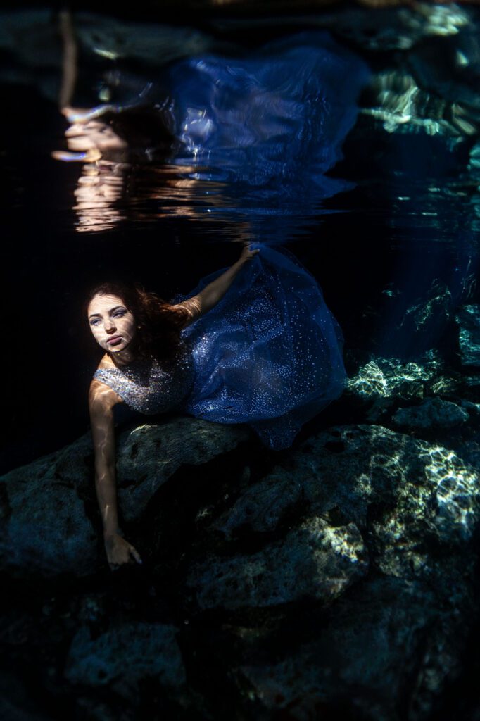 A girl poses underwater with her shiny dress. She is lying face down on a rock and looking straight into the camera.